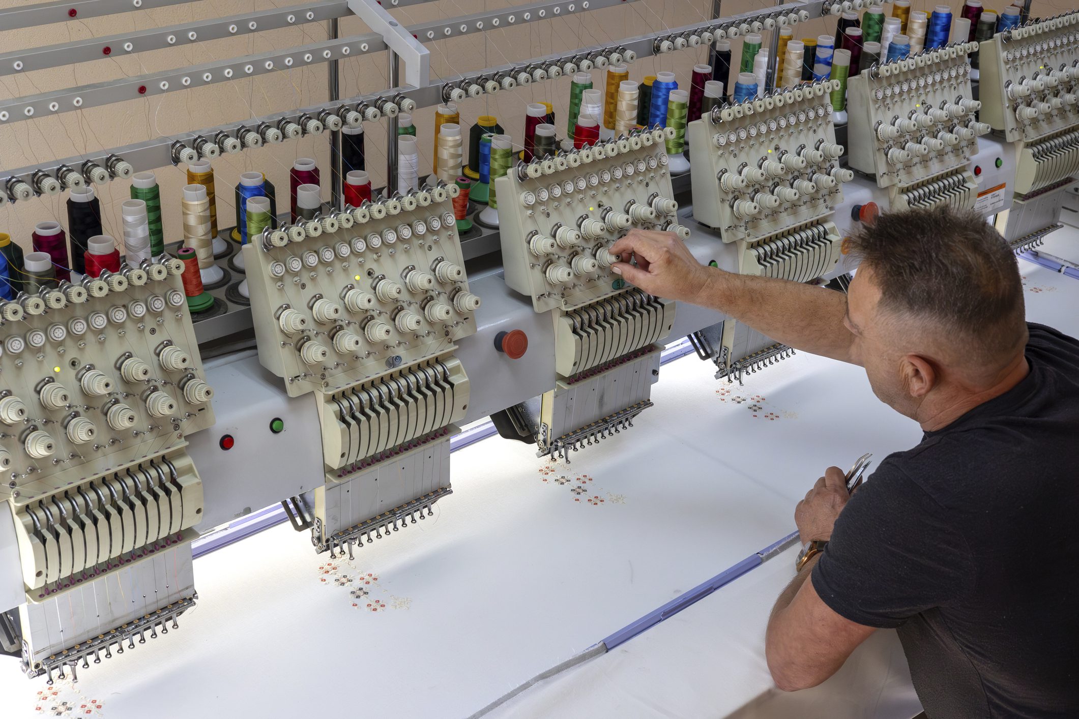 Male craftsman supervising embroidery process on large industrial embroidery machine in sewing workshop. Crafts, small business, fashion branding, textile manufacturing and embroidery art topics. Man operating industrial embroidery machine.